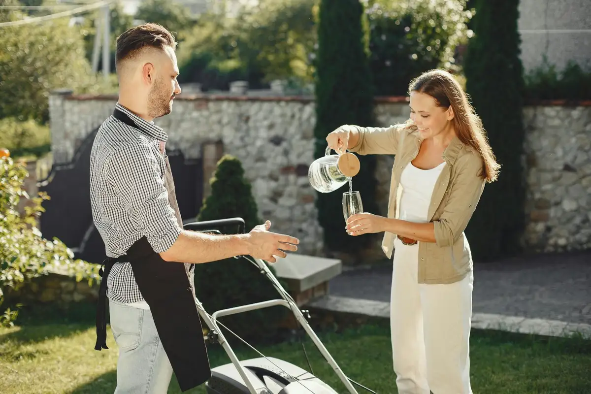 Two adults outdoors beside a lawn mower; a man wearing a checkered shirt and apron discusses maintenance with a woman who holds a pitcher.