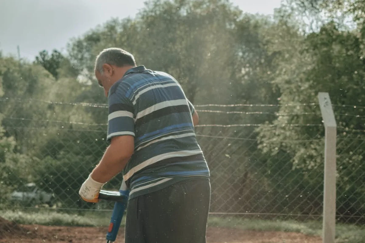 Older man wearing a striped polo shirt mowing a dry lawn beside a chain-link fence.