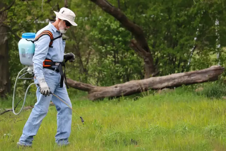 Lawn care professional wearing protective gear and a backpack sprayer walks across a grassy field with a fallen tree in the background.