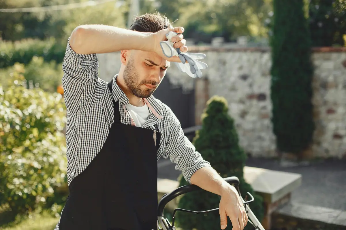 Man in a checkered shirt wearing a black apron adjusts the lawn mower's blade height outdoors using a tool.