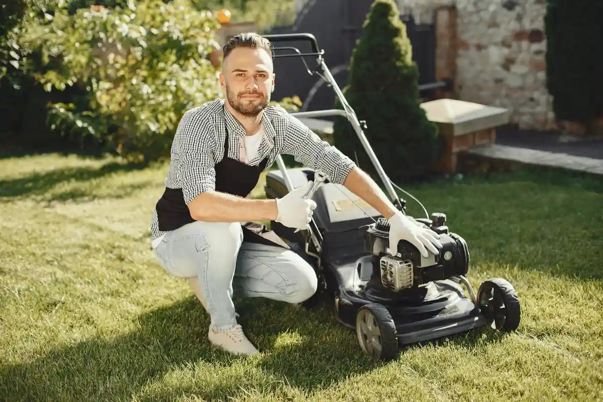 Man kneeling on a sunny lawn beside a gas-powered lawn mower, inspecting it before jump-starting the battery.