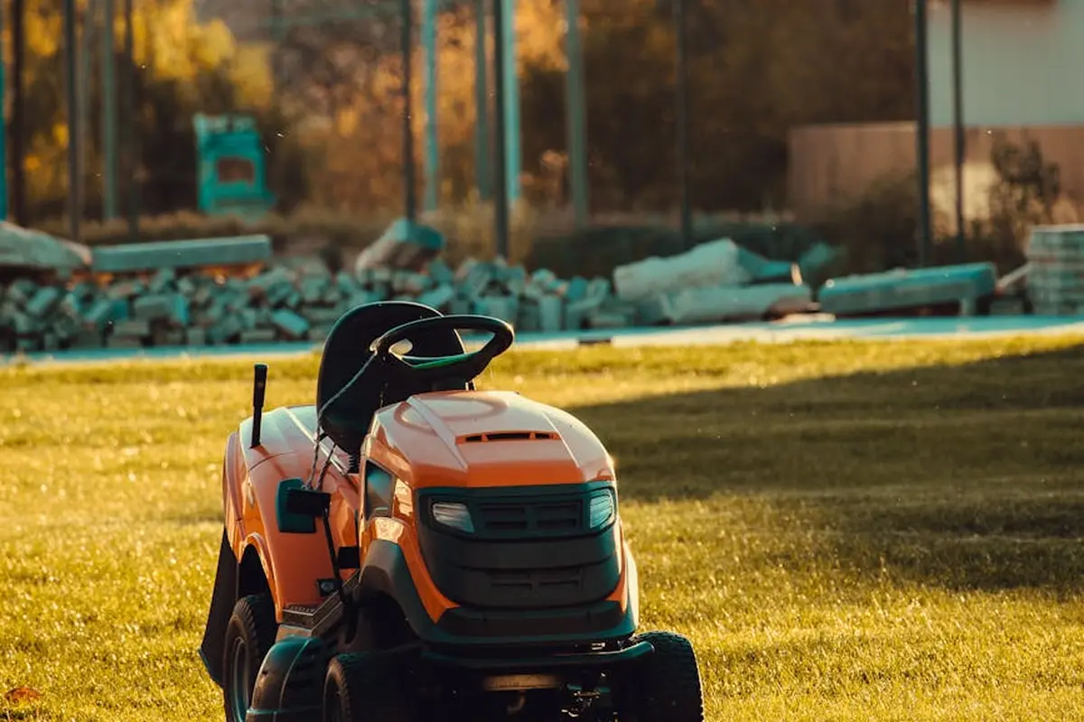Orange riding lawn mower resting on a sunlit lawn with a blurred construction site in the background.