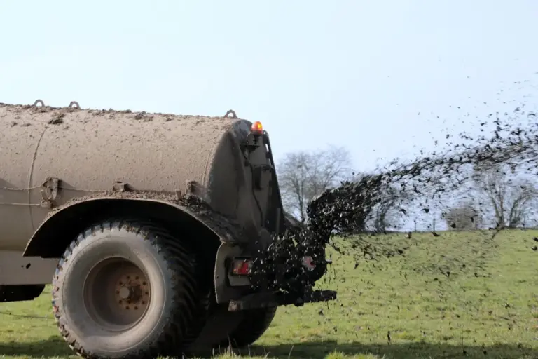 A large tanker sprayer applying liquid fertilizer to a grassy field, spraying a mist behind as it travels.