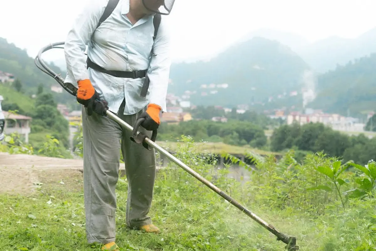 Person wearing gloves and long-sleeve shirt using a string trimmer in a grassy field outdoors.