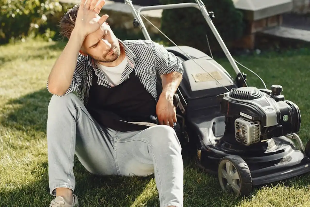 A man sits on the grass beside a lawn mower, shading his eyes with one hand.