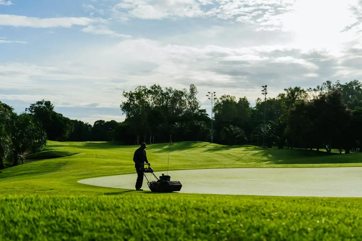 A groundskeeper pushes a manual aeration tool across a well-manicured golf course green.