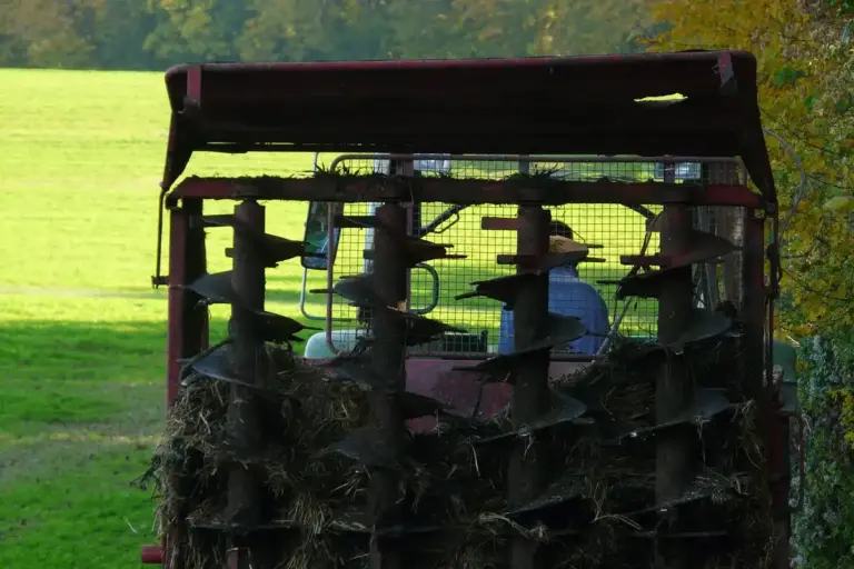 Red manure spreader filled with organic matter in a sunlit grassy field.