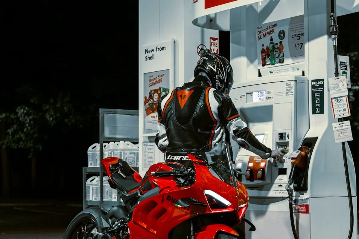 A person in motorcycle riding gear stands beside a red sport bike refueling at a gas pump.
