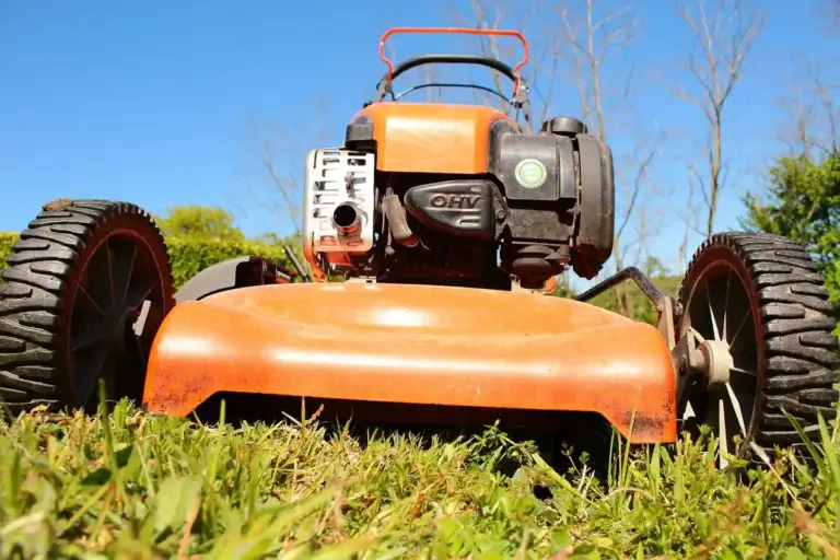 Low-angle close-up of an orange gas-powered lawn mower resting on a grassy lawn with large wheels and blue sky in the background.