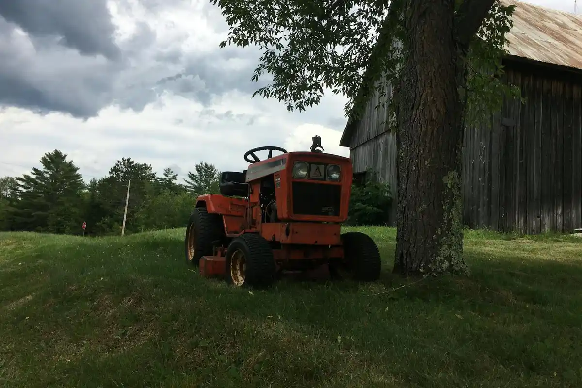 An orange lawn tractor parked on a grassy slope beside a weathered wooden barn under a cloudy sky.