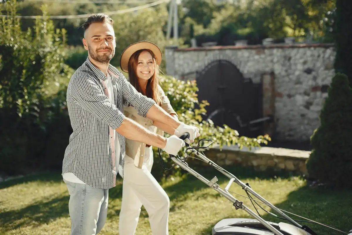 Smiling couple in a sunny backyard pushing a lawn mower, illustrating everyday lawn care as part of planning dethatching.