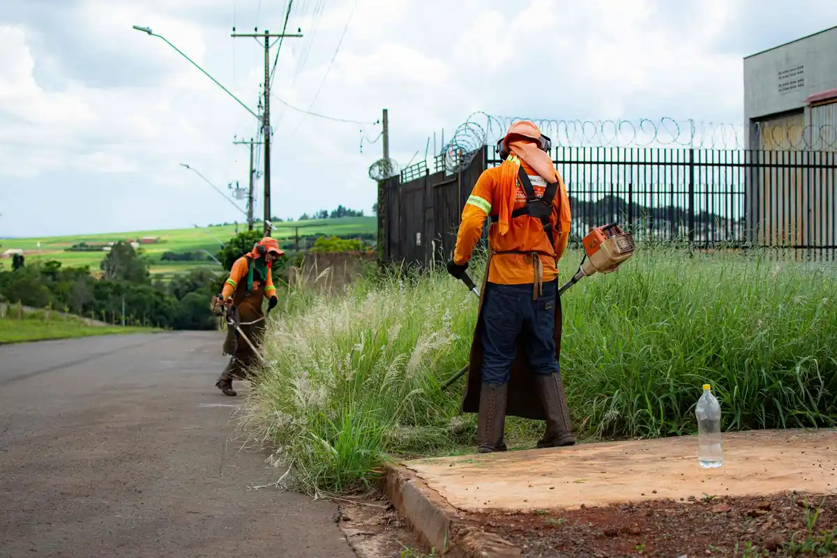 Two workers in orange safety shirts mowing tall grass along a roadside, with utility poles and a chain-link fence in the background.