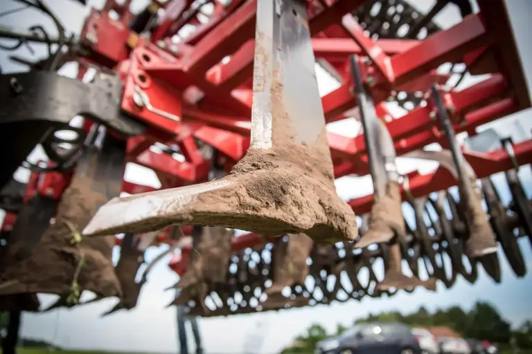 Close-up of a seed drill's metal tines with dirt on them and a red framework in the background.