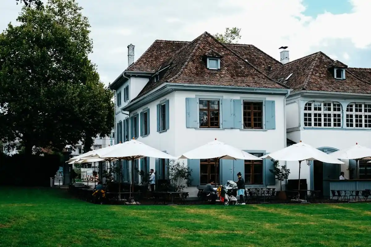 Exterior of a large white house with blue shutters and a patio with white umbrellas on a green lawn.