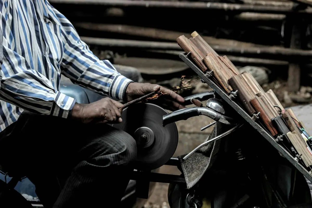 A person wearing a striped shirt sharpens a lawn mower blade on a bench grinder, with blades clamped in a vise nearby.