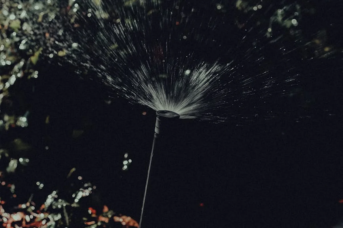 A lawn sprinkler emits a spray arc across a dark garden, illustrating how numerous spray patterns cover different areas