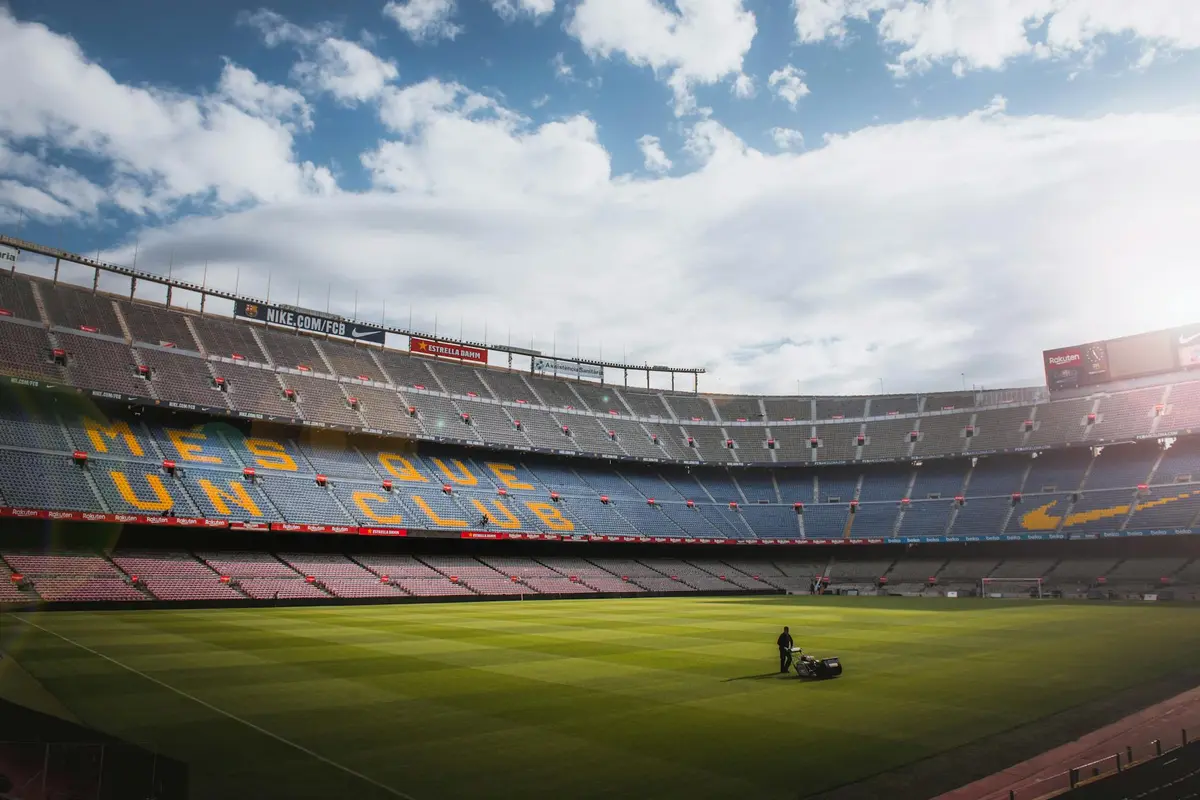 A person mowing the expansive grass on a large stadium field with empty stands and a bright sky