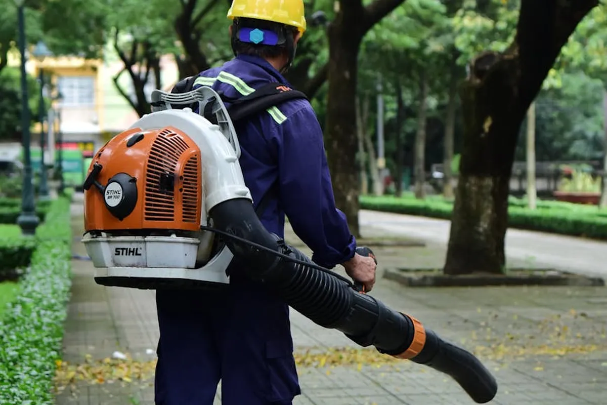 Groundskeeper wearing a safety helmet and protective gear, using a Stihl backpack leaf blower on a lawn path