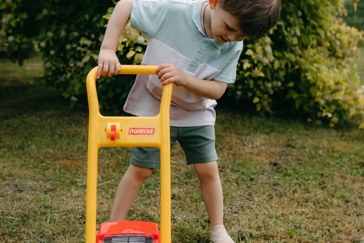 A child pushes a yellow toy lawn mower across a grassy yard.
