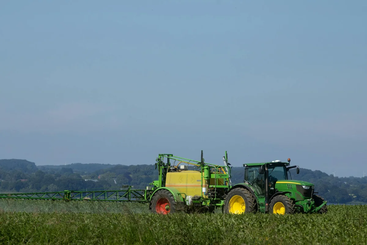 Green tractor with a yellow-wheeled fertilizer spreader in a grassy field under a clear blue sky.