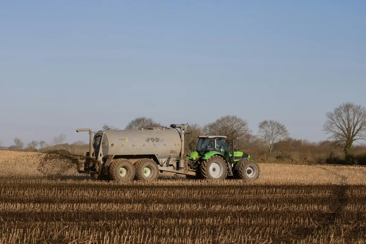 Green tractor towing a large gray fertilizer tanker, applying fertilizer across a plowed berry field.