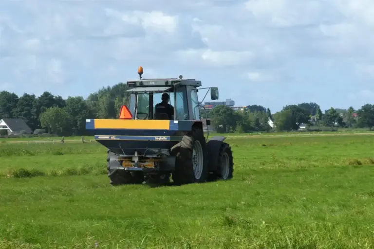 Agricultural tractor with a fertilizer spreader in a bright green field under a blue sky.