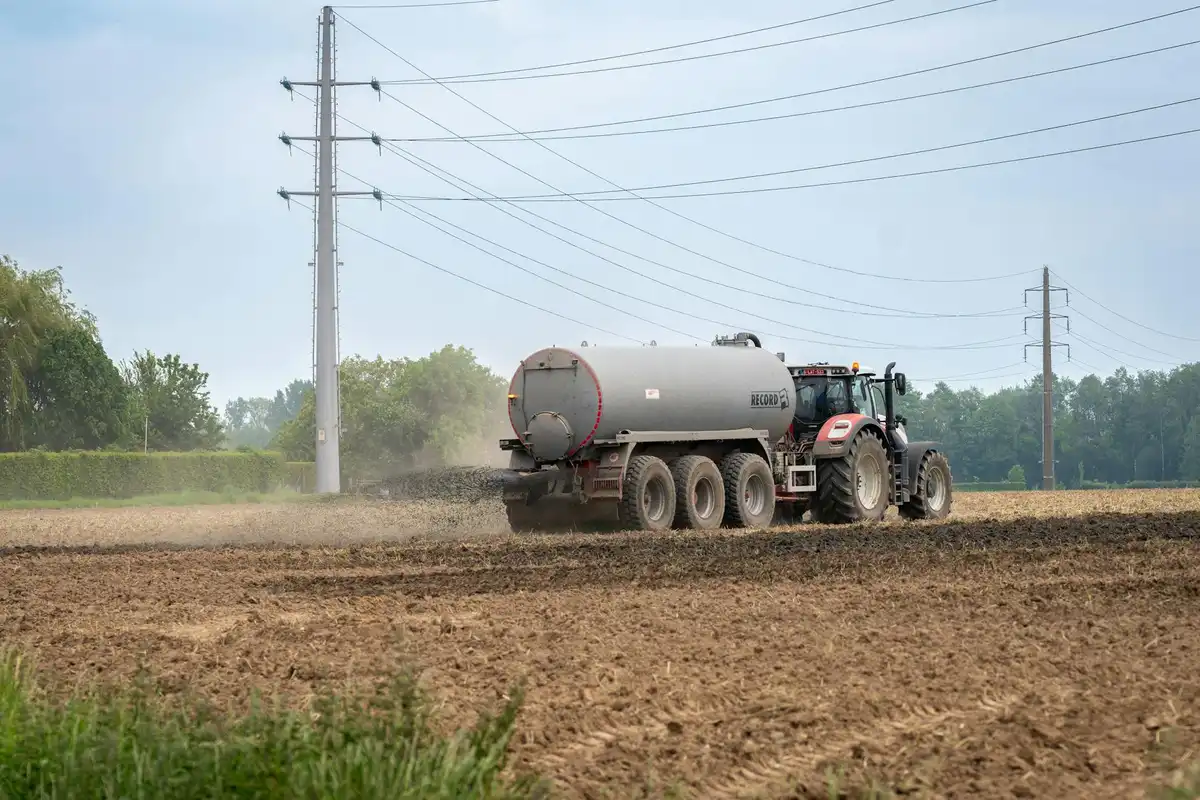 A tractor with a large fertilizer spreader moves across a tilled field under utility poles, applying nutrients to the soil.
