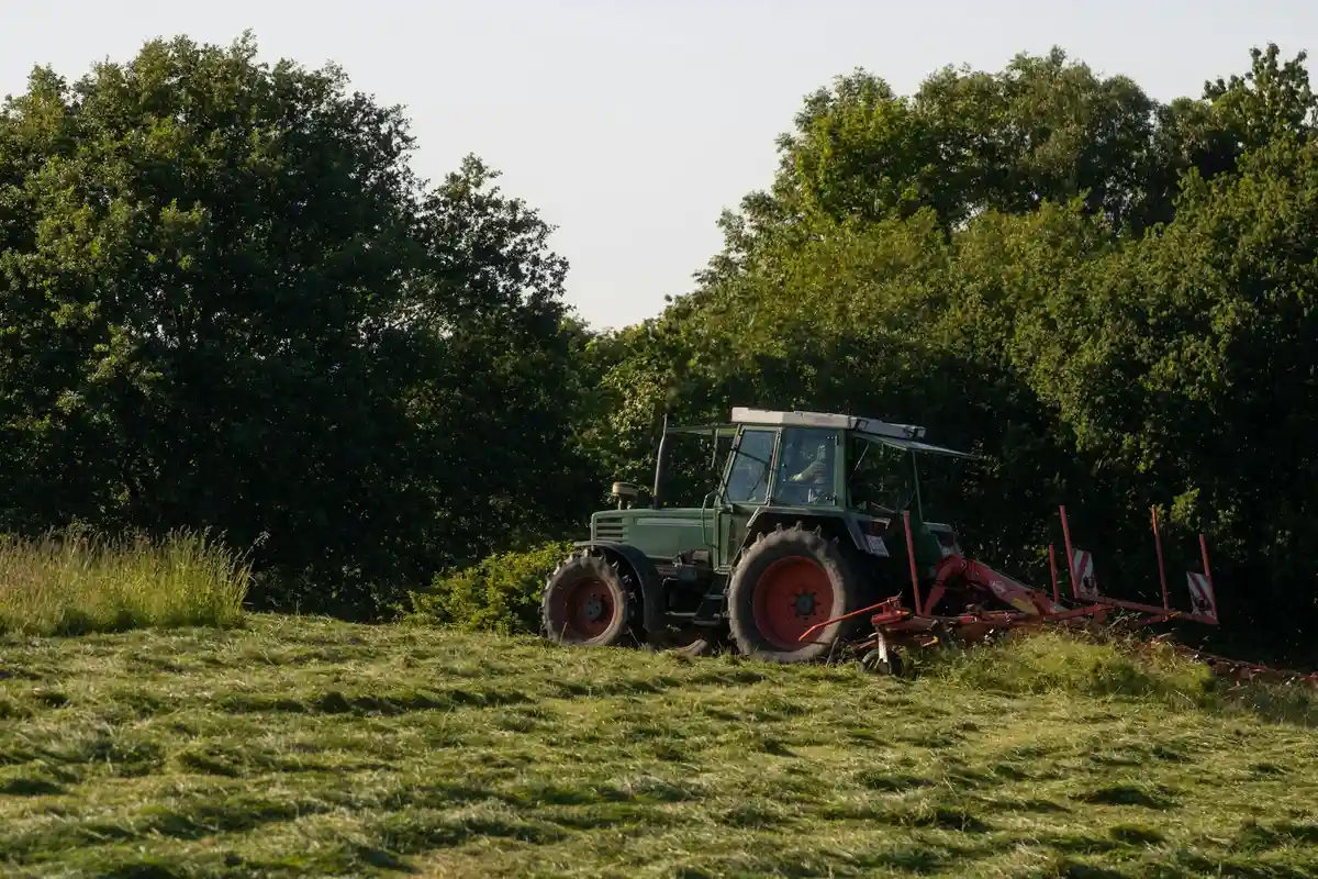 Green tractor with a red mowing attachment sits on a grassy field with trees in the background under a sunny sky.