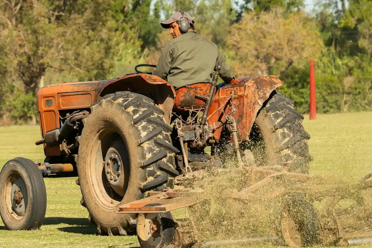 A person driving an old tractor with a mower attachment cutting through an overgrown lawn, kicking up grass clippings and dust.