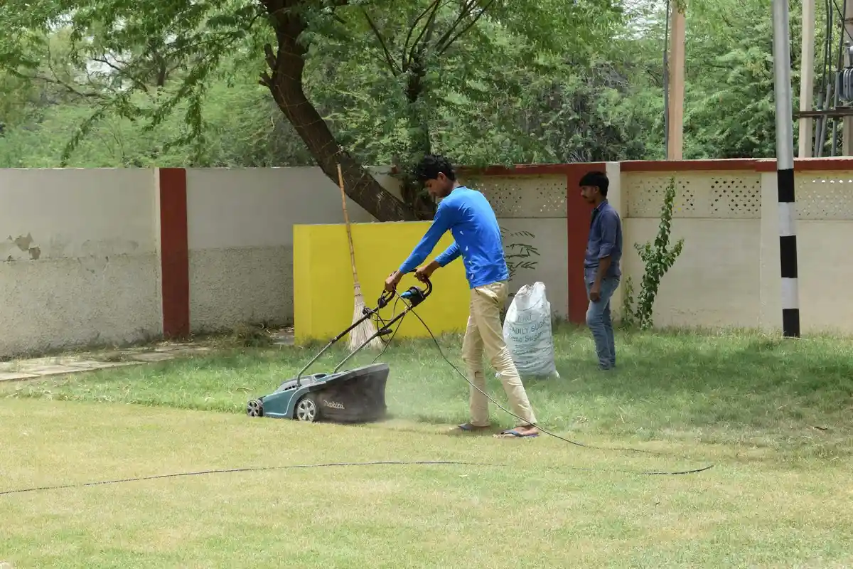 Two men mowing a lawn with a push-style lawn mower in a backyard, one actively pushing the mower while the other watches.