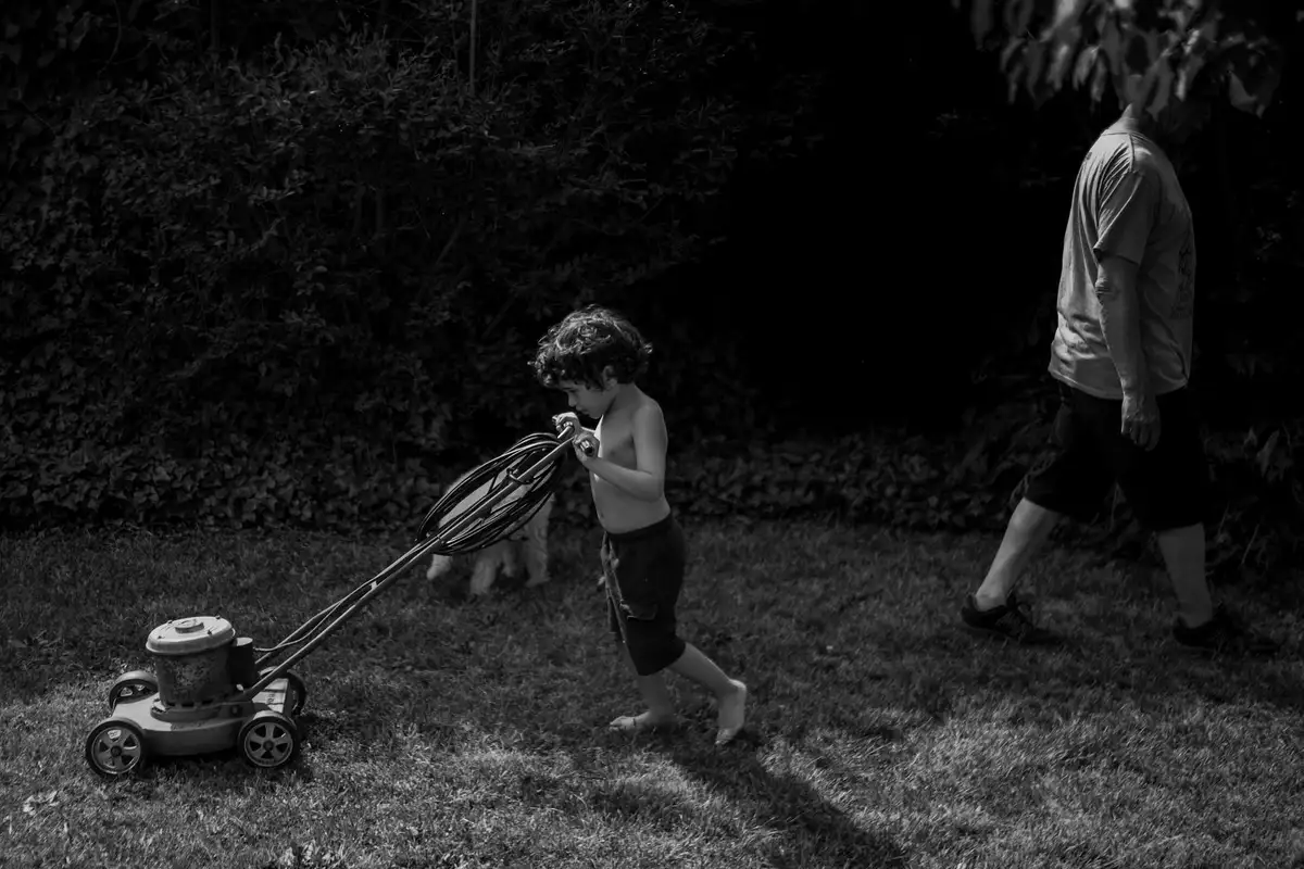 A child pulls a push lawn mower across a wet lawn while an adult walks nearby.