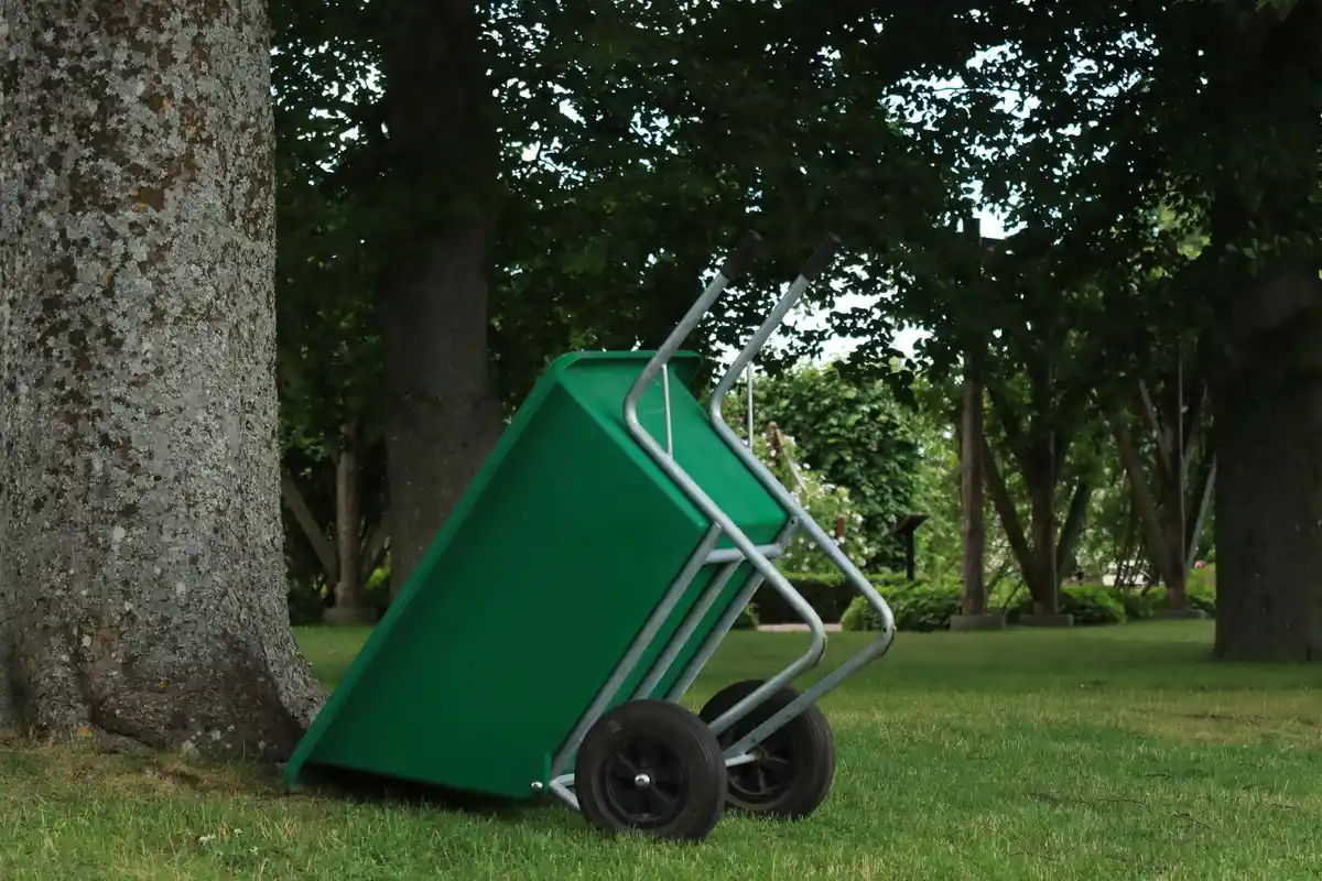 A green wheelbarrow resting on a grassy lawn beside several trees in a park.