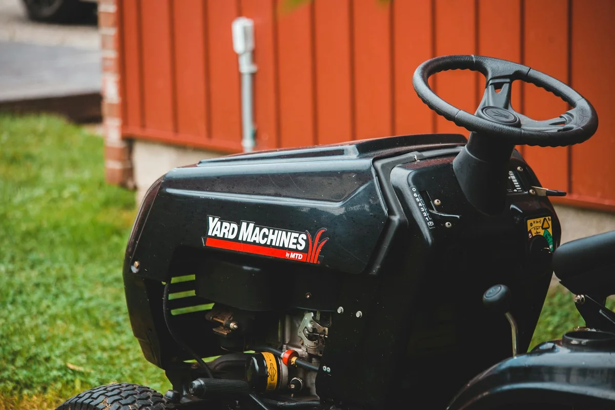 Black Yard Machines riding lawn mower with steering wheel, parked on grass beside a red fence