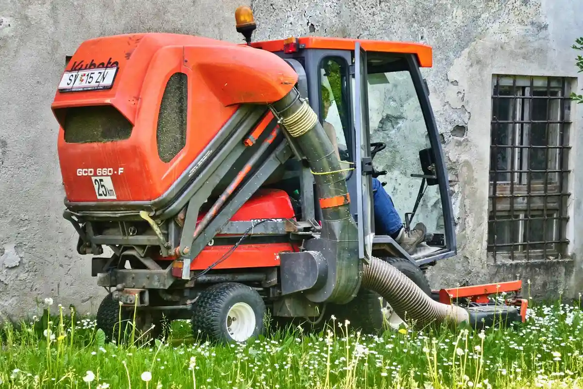 Red ride-on lawn mower with a bagging attachment, parked on a grassy area beside a weathered wall with a window