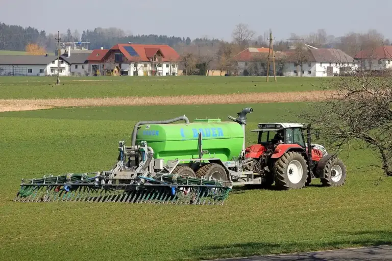 Red tractor towing a large green fertilizer sprayer across a grassy field with residential houses in the background.