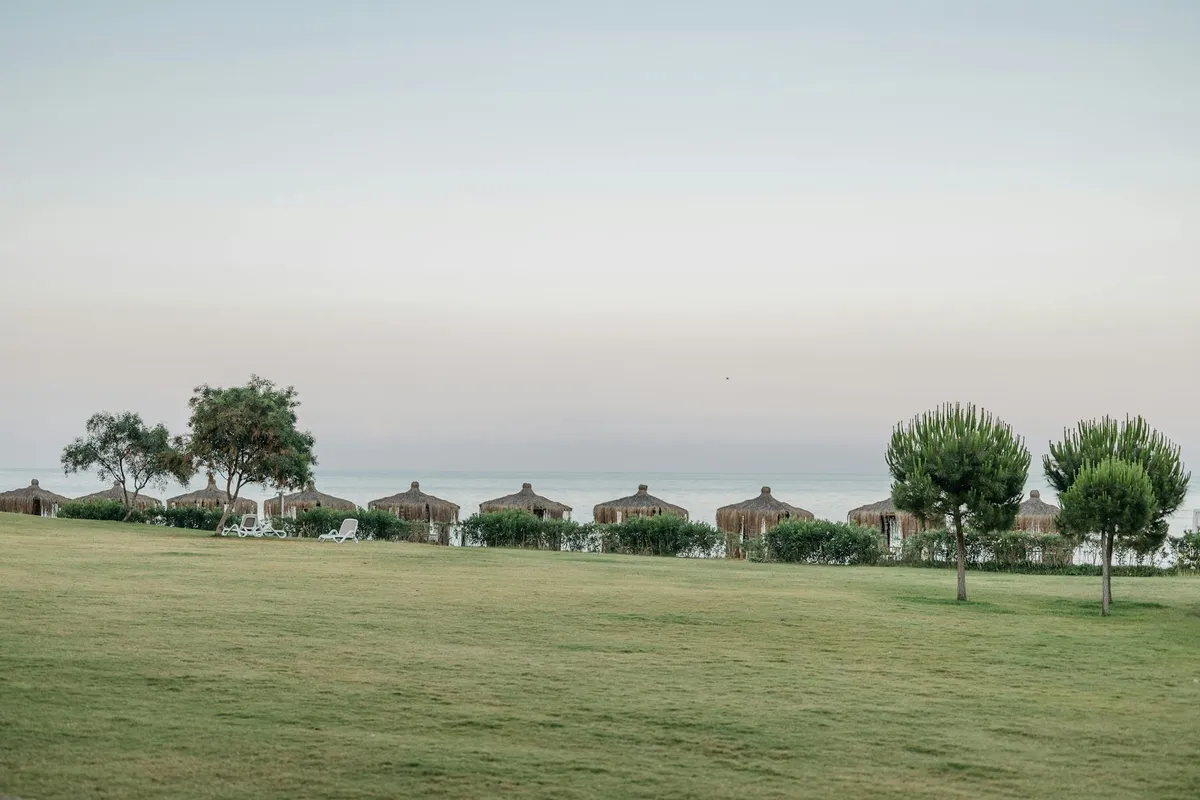 Wide grassy field with distant seaside cottages along the horizon and a few trees.