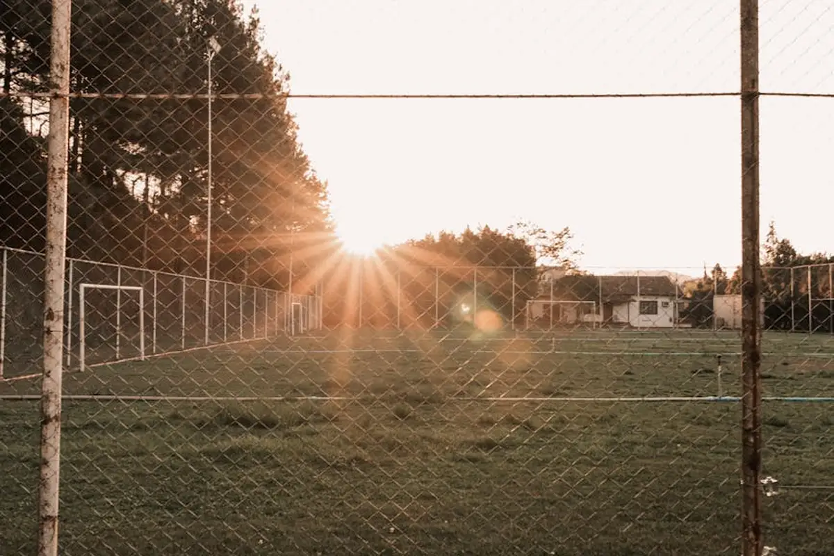 Chain-link fence surrounding a grassy sports field at sunset, with the sun visible through the fence.
